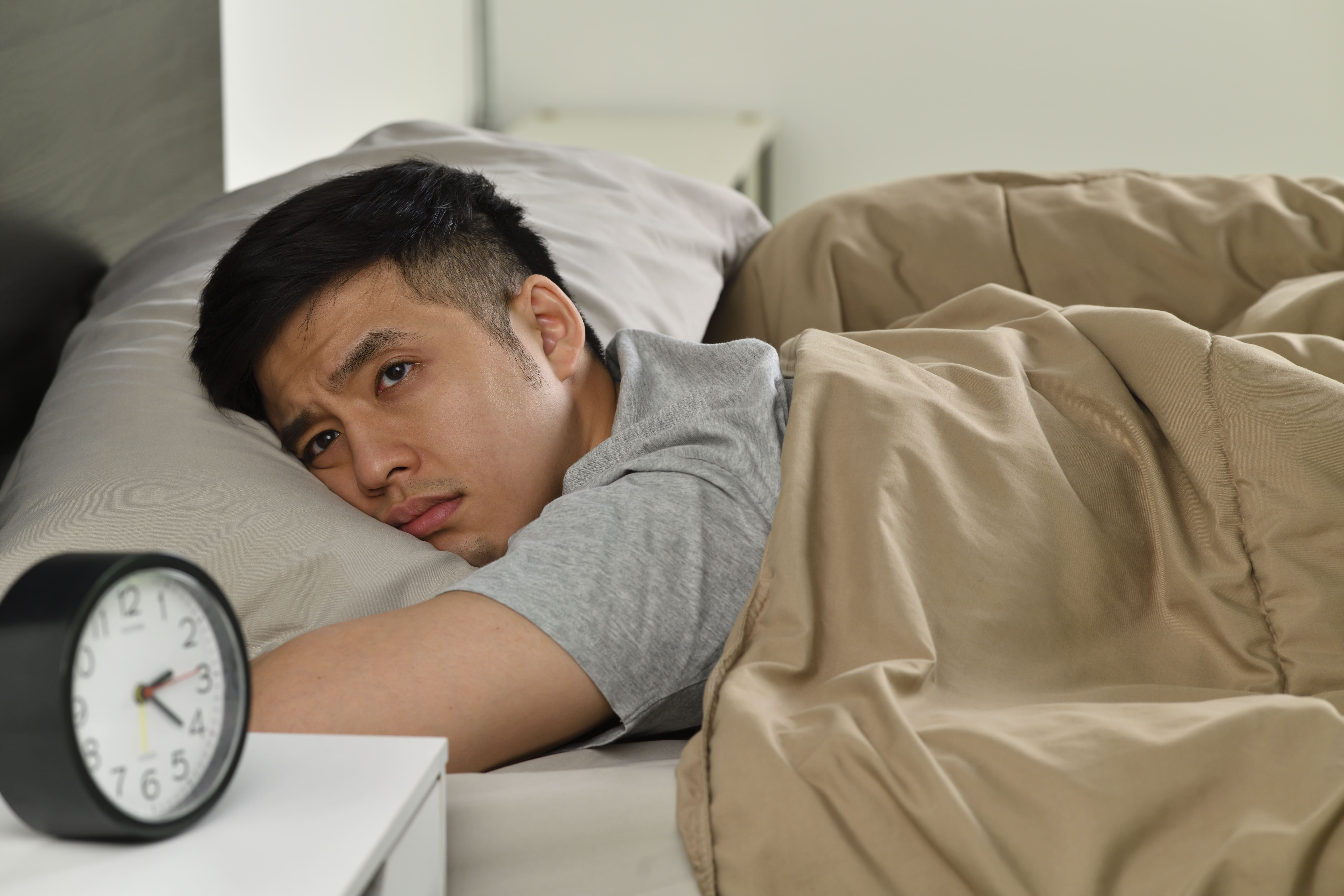 A person lying in bed under a blanket next to an alarm clock, appearing awake.